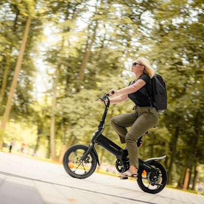 Na sliki je električno kolo, model MS Energy Urbanfold i6. Zasnova kolesa je kompaktna in zložljiva, kar omogoča enostavno prenašanje. Na kolesu vozi ženska, oblečena v črne majice in olivno zelene hlače, ki nosi nahrbtnik. Ozadje prikazuje zeleno okolje, kar nakazuje, da se kolo uporablja v naravi ali parku.