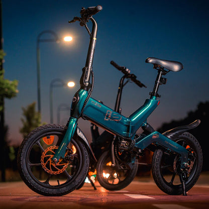 Izdelek na sliki je električno kolo, model MS Energy Urbanfold i6. Kolo je kompaktne zasnove z manjšimi kolesi in preklopnim mehanizmom, kar omogoča enostavno shranjevanje in prenašanje. Barva okvirja je svetlo modra, na sprednjem kolesu pa so vidne disk zavore. V ozadju so vidne ulične svetilke, kar ustvarja prijetno vzdušje v večernih urah.