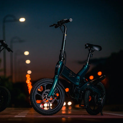 Izdelek na sliki je električno kolo, model MS Energy Urbanfold i6. Kolo ima kompakten in zložen dizajn, kar omogoča enostavno shranjevanje in prenašanje. Na sliki je vidna temno zelena barva okvirja, široka kolesa ter ergonomična sedežna in krmilna pozicija, kar nakazuje na udobno vožnjo. Osvetlitev v ozadju ustvarja prijetno nočno atmosfero.