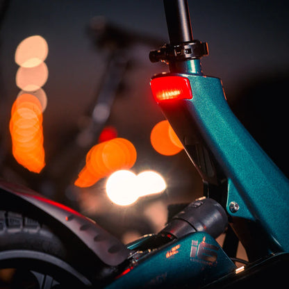 Izdelek na sliki je električno kolo, model MS Energy Urbanfold i6. Na sliki je vidna podrobnost okvirja kolesa v temno zeleni barvi, s poudarjenim zadnjim lučjo, ki sveti rdeče. Osvetlitev in bokeh učinek ozadja ustvarjata dinamično nočno vzdušje, kar nakazuje na uporabo kolesa v urbanem okolju.