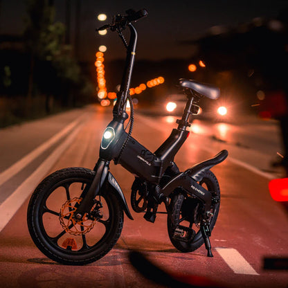 Izdelek na sliki je električno kolo, model MS Energy Urbanfold i6. Kolo je kompaktne in moderne zasnove, opremljeno z LED lučjo spredaj, kar omogoča vožnjo tudi v slabših svetlobnih pogojih. Ima široke gume in zložljivo konstrukcijo, kar ga naredi primernega za mestno uporabo in enostavno shranjevanje. Na sliki je vidna urbana okolica, kar dodatno poudarja njegovo uporabnost v mestnem prometu.