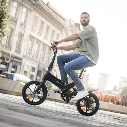 Izdelek na sliki je električno kolo, znamke MS Energy, model Urbanfold i6. Na sliki je moški, ki sedi na kolesu, oblečen v svetlo sivo majico in modre kavbojke. Kolo ima kompaktno zasnovo in je primerno za urbano vožnjo, kar je vidno tudi v ozadju, kjer so mestne zgradbe. Kolo ima majhne kolesa in je opremljeno z dodatnimi varnostnimi elementi, kot so odsevniki.