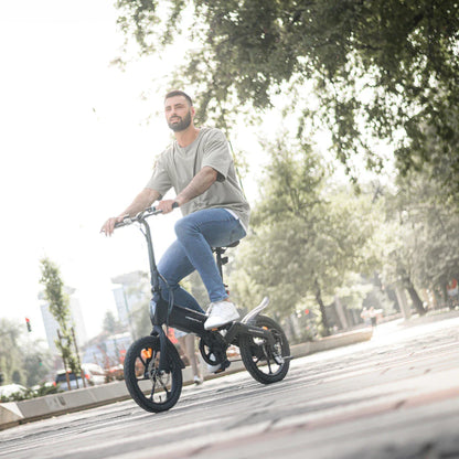 Na sliki je električno kolo, model MS Energy Urbanfold i6. Kolo ima kompakten dizajn in je primerno za mestno vožnjo. Na sliki je moški, ki sedi na kolesu in se vozi po urejeni mestni površini, obdani z drevesi in zelenjem. Kolo je opremljeno z manjšimi kolesi, kar omogoča enostavno manevriranje v urbanem okolju.