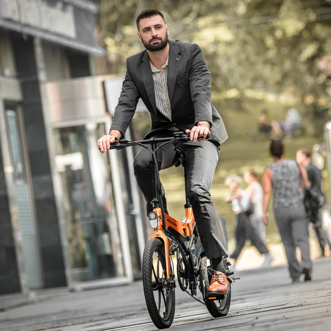 Na sliki je električno kolo, model MS Energy i20. Kolo je vidno v urbanem okolju, kjer moški v poslovnem obleku kolesari. Ima svetlo oranžno barvo in je zasnovano za udobno vožnjo po mestnih ulicah. Opremljeno je z manjšimi kolesi in športnim dizajnom, kar omogoča enostavno manevriranje. V ozadju so vidni tudi drugi ljudje, kar nakazuje na živahno mestno okolje.