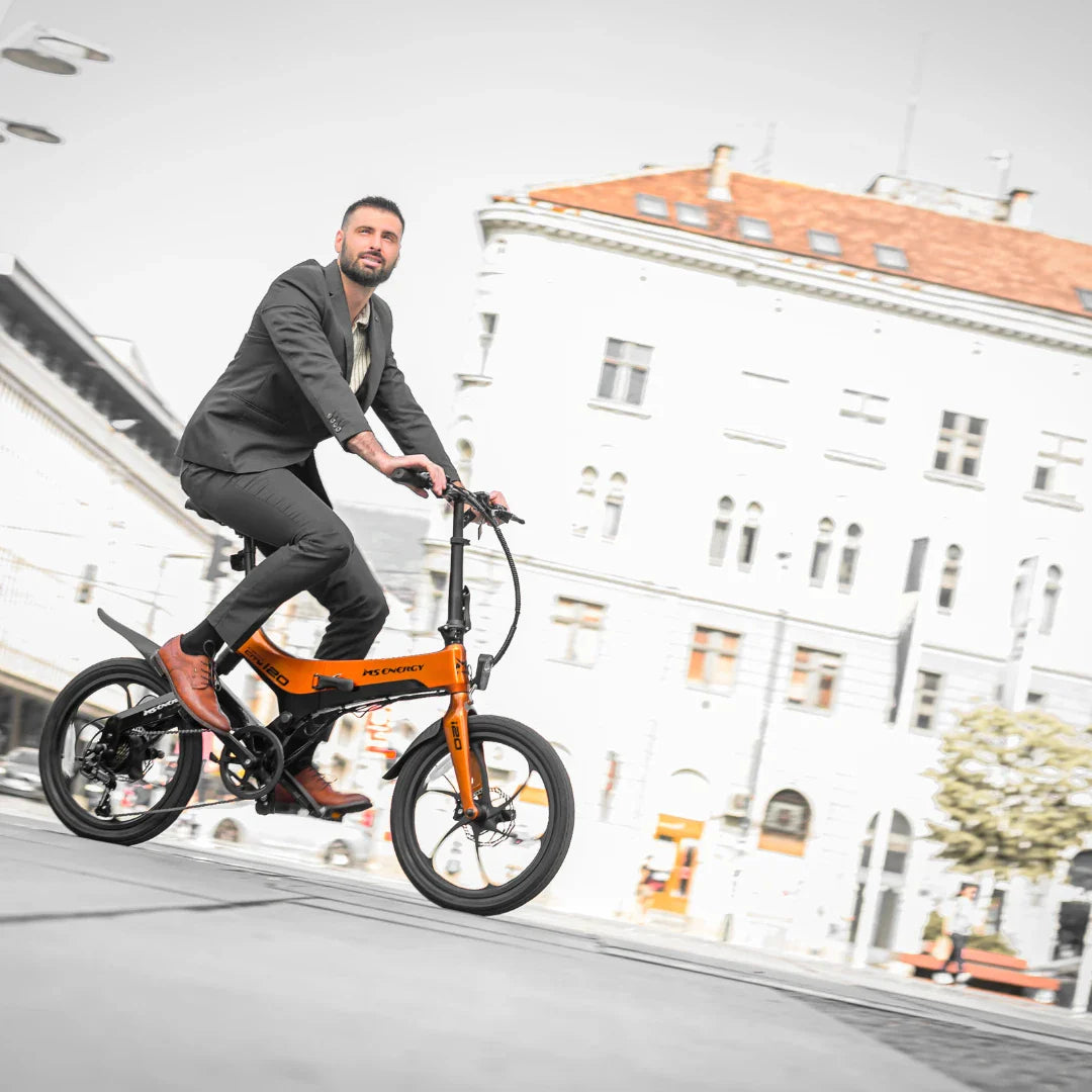 Na sliki je električno kolo, model MS Energy I20. Kolo je v svetlo oranžni barvi in ima sodoben dizajn. Na njem je moški oblečen v poslovno obleko, ki se vozi po mestni ulici. V ozadju so vidne mestne zgradbe, kar nakazuje urbano okolje. Električno kolo je primerno za udobno in hitro premikanje po mestu.