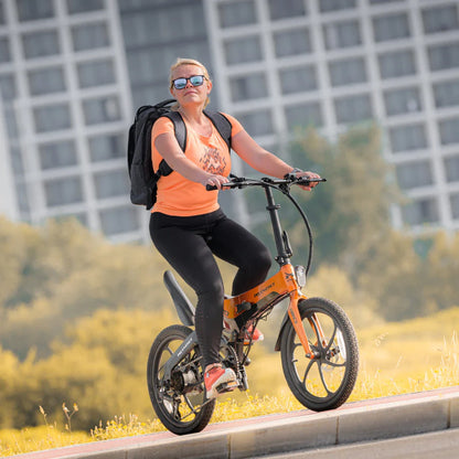 Izdelek na sliki je električno kolo, model MS Energy i20. Na sliki vidimo voznico, ki se vozi s kolesom po poti, oblečeno v športna oblačila in s nahrbtnikom na hrbtu. Kolo je v svetlo oranžni barvi in ima zložljivo zasnovo, kar ga naredi praktičnega za prevoz in shranjevanje. V ozadju so vidne zgradbe in narava, kar nakazuje urbano okolje.