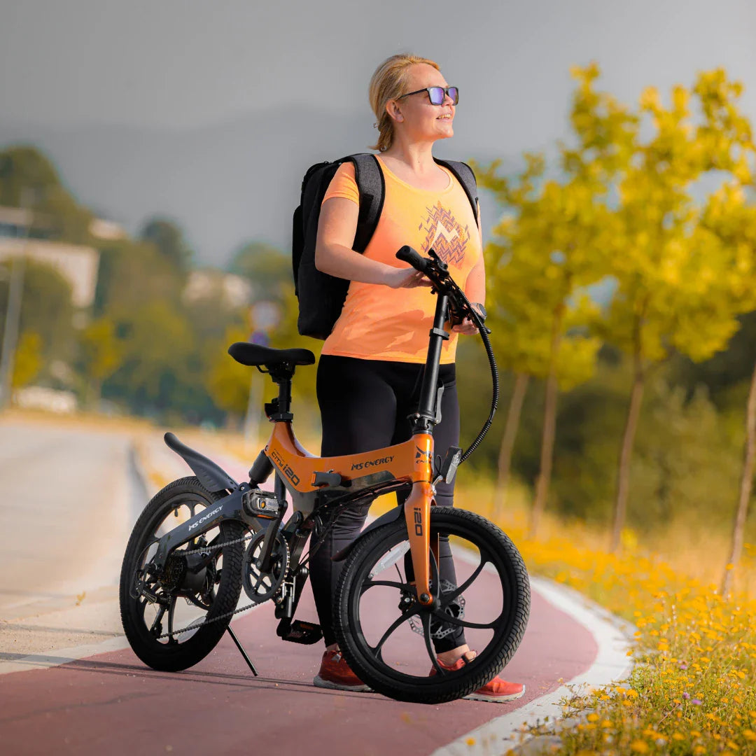 Na sliki je električno kolo znamke MS Energy, model i20. Prikazuje mlado žensko osebo, ki stoji ob kolesu na poti, oblečeno v športno majico in črne hlače, ter nosi nahrbtnik. Kolo ima svetlo oranžno barvo in je zasnovano za udobno vožnjo. V ozadju so drevesa in narava, kar ustvarja prijeten ambient za kolesarjenje.