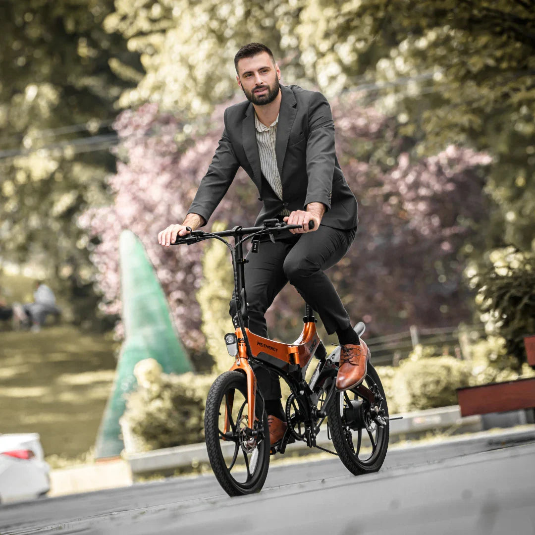 Izdelek je električno kolo, model MS Energy i20. Na sliki je moški v poslovnem obleku, ki vozi to električno kolo po urejeni poti v parku. Kolo ima moderno obliko in je v oranžni barvi, kar ga naredi privlačnega za uporabnike. V ozadju so drevesa in zelenje, kar daje sliki prijeten ambient.