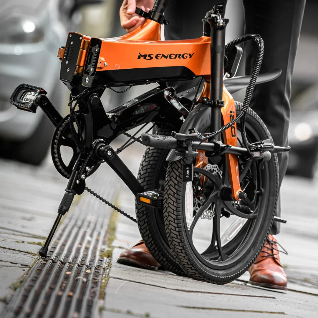 Izdelek na sliki je električno kolo znamke MS Energy, model i20. Kolo je zložljivo in opremljeno s sodobnim dizajnom ter robustnimi pnevmatikami. Na sliki je vidna oseba, ki drži kolo, medtem ko je eno kolo na tleh. Kolo je postavljeno na stabilnem stojalu, kar nakazuje, da je pripravljeno za uporabo ali shranjevanje.