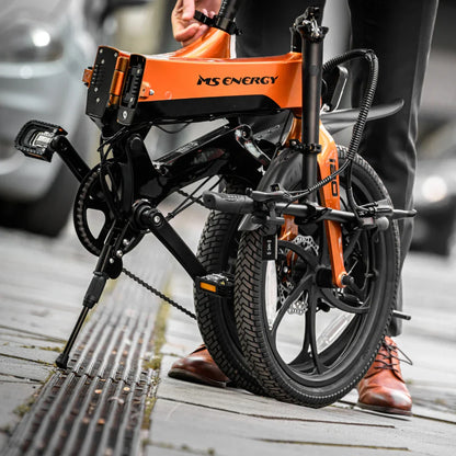 Izdelek na sliki je električno kolo znamke MS Energy, model i20. Kolo je zložljivo in opremljeno s sodobnim dizajnom ter robustnimi pnevmatikami. Na sliki je vidna oseba, ki drži kolo, medtem ko je eno kolo na tleh. Kolo je postavljeno na stabilnem stojalu, kar nakazuje, da je pripravljeno za uporabo ali shranjevanje.