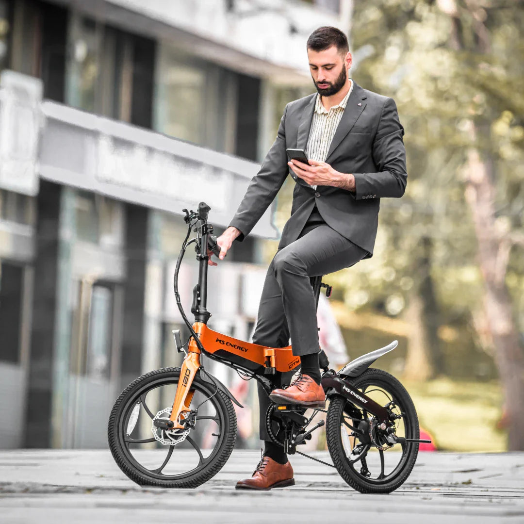 Na sliki je električno kolo, model MS Energy I20. Zasnova kolesa je kompaktna in moderna, kar omogoča enostavno vožnjo po mestnih ulicah. Moški v poslovnem obleku sedi na kolesu in uporablja mobilni telefon, kar kaže na njegovo praktičnost in primernost za urbano okolje. Kolo ima značilne manjše kolesa in športen videz, kar je značilno za električna kolesa te vrste.