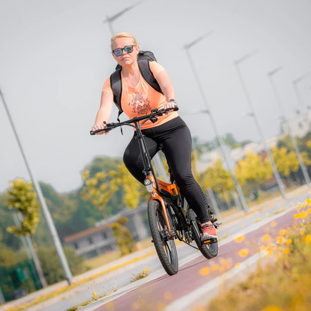 Na sliki je električno kolo MS Energy I20. Vidimo žensko, ki vozi kolo po urejeni poti, oblečeno v športno oblačilo in s nahrbtnikom na hrbtu. Kolo je v svetli oranžni barvi in ima športen videz, kar nakazuje na njegovo uporabnost za aktivno prevozno sredstvo. V ozadju so drevesa in urbana okolica.