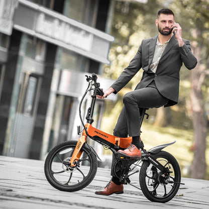 Izdelek na sliki je električno kolo znamke MS Energy, model I20. Na sliki je moški v poslovnem obleku, ki sedi na električnem kolesu in govori po telefonu. Kolo ima kompaktno zasnovo, značilno za mestna prevozna sredstva, in je v svetlo oranžni barvi. V ozadju so vidni urbani elementi, kar nakazuje, da je kolo primerno za mestno vožnjo.