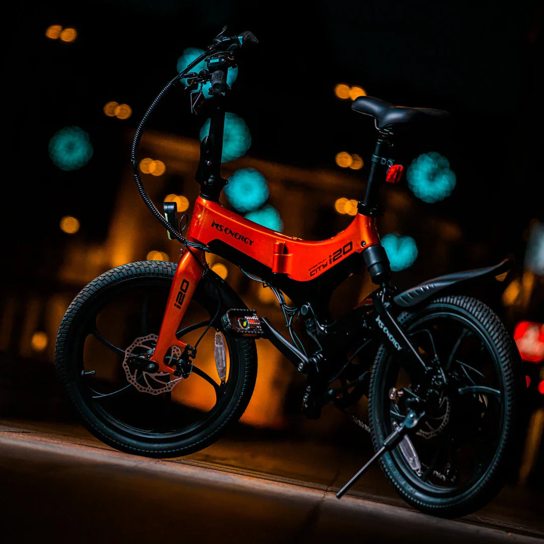 Izdelek na sliki je električno kolo znamke MS Energy, model i20. Kolo ima značilno svetlo oranžno barvo s črnimi detajli in je opremljeno z debelimi pnevmatikami ter modernim dizajnom. Na sliki je kolo postavljeno v nočnem okolju, kar poudarja njegove oblikovne linije in detajle.