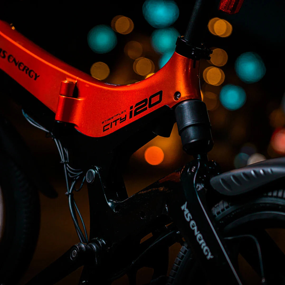 Izdelek na sliki je električno kolo, znamke MS Energy, model i20. Na sliki je prikazan del okvirja v svetlo rdeči barvi, ki je opremljen s črnim delom, kjer se nahajajo kolesa in mehanizem. V ozadju so zamegljene luči, kar daje sliki nočno vzdušje.