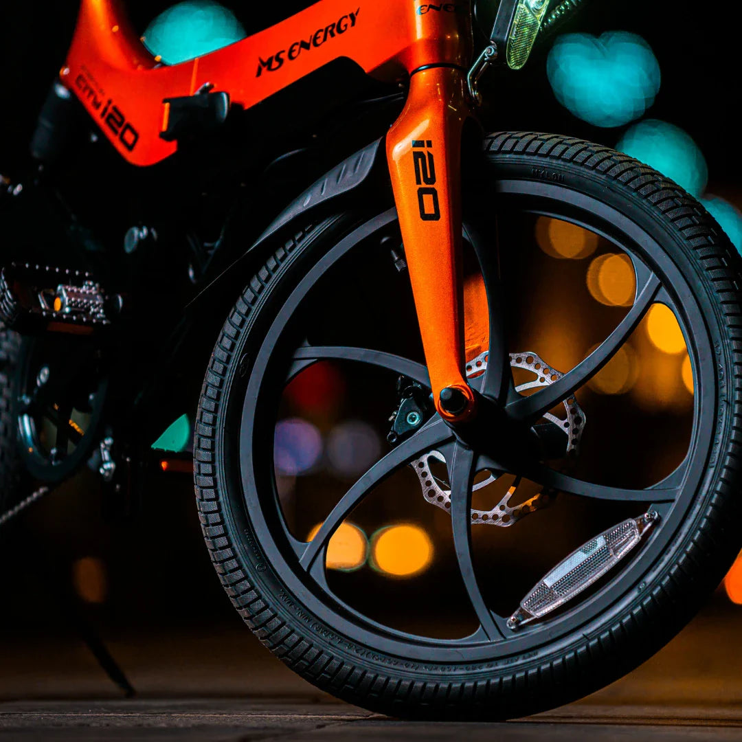Na sliki je električno kolo znamke MS Energy, model i20. Vidna je sprednja vilica in kolo, ki ima sodoben dizajn z več kraki in disk zavornim sistemom. Barva okvirja je svetlo oranžna, kar daje kolesu dinamičen videz. V ozadju so mehki bokeh učinki, kar ustvarja prijetno nočno atmosfero.