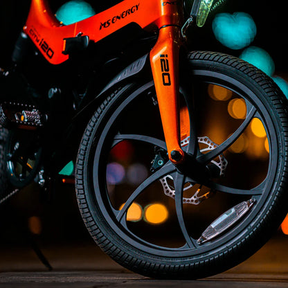 Na sliki je električno kolo znamke MS Energy, model i20. Vidna je sprednja vilica in kolo, ki ima sodoben dizajn z več kraki in disk zavornim sistemom. Barva okvirja je svetlo oranžna, kar daje kolesu dinamičen videz. V ozadju so mehki bokeh učinki, kar ustvarja prijetno nočno atmosfero.
