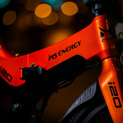 Izdelek na sliki je električno kolo znamke MS Energy, model i20. Kolo ima svetlo oranžno barvo in je opremljeno z modernim okvirjem. Na sliki so vidni detajli okvirja, ki vključujejo logotip znamke in oznako modela. Osvetlitev v ozadju dodaja dinamičen občutek, kar poudarja sodobno zasnovo kolesa.