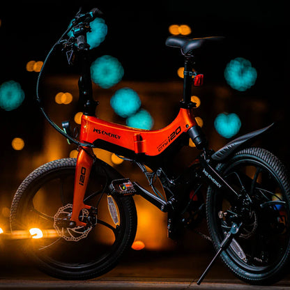 Izdelek na sliki je električno kolo, znamke MS Energy, model i20. Kolo ima svetlo oranžno barvo okvirja in je zasnovano za mestno vožnjo. Na sliki je vidna tudi njegova konstrukcija, vključno s kolesi, krmilom in sedežem, ter ozadje, ki ustvarja prijetno nočno vzdušje z zamegljenimi svetlobnimi točkami.