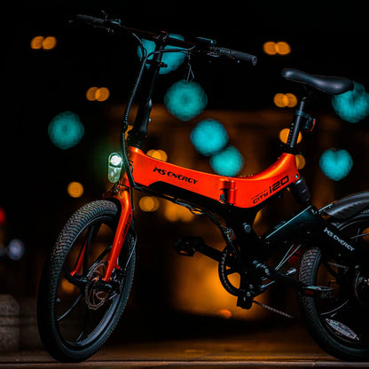 Izdelek na sliki je električno kolo, znamke MS Energy, model i20. Kolo je v svetlo oranžni barvi, opremljeno z LED lučjo na sprednjem delu in ima kompaktno zasnovo, kar ga naredi primerno za mestno vožnjo. V ozadju so mehki svetlobni učinki, kar daje sliki dinamičen videz.