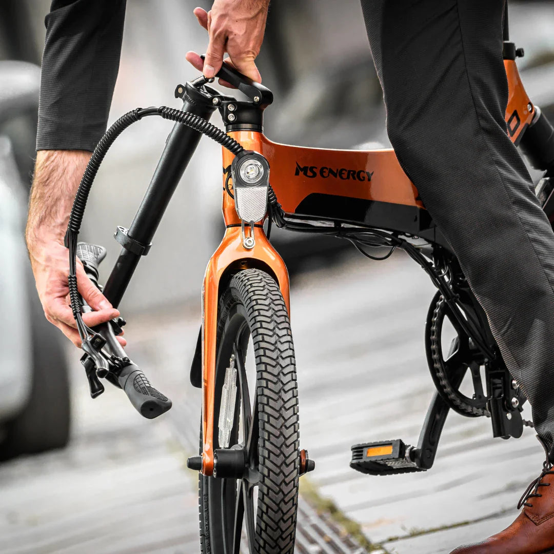 Izdelek na sliki je električno kolo, znamke MS Energy, model i20. Na sliki je vidna oseba, ki drži del kolesa, verjetno krmilo, in se pripravlja na vožnjo. Kolo ima značilno obliko z debelimi pnevmatikami in sodobnim dizajnom. Barva okvirja je oranžna, kar mu daje privlačen videz.