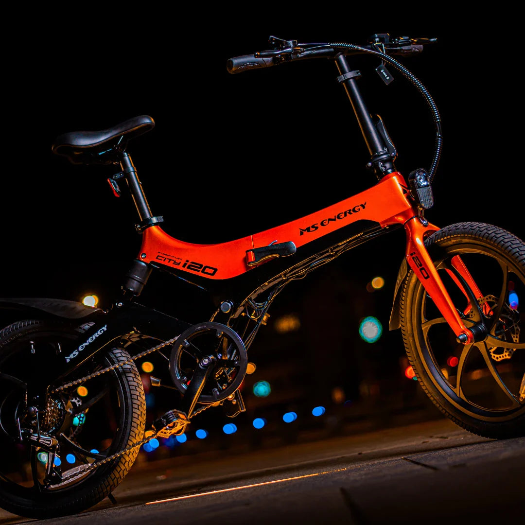 Izdelek na sliki je električno kolo, znamke MS Energy, model i20. Kolo ima kompaktno zasnovo, s črno in rdečo barvno kombinacijo ter športnim videzom. Vidni so tudi detajli, kot so krmilo, sedež in kolesa, kar nakazuje na njegovo uporabnost v urbanem okolju. Osvetlitev v ozadju dodaja dinamičnost in modernost celotni podobi.
