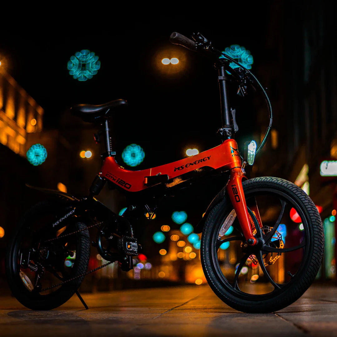 Izdelek na sliki je električno kolo znamke MS Energy, model i20. Kolo ima kompakten dizajn in je v živahni rdeči barvi. Na sliki je vidno tudi nočno okolje z raznobarvnimi lučmi v ozadju, kar daje kolesu dinamičen videz. Kolo je opremljeno z debelimi pnevmatikami in ima klasične krmilo ter sedež, kar nakazuje na udobno vožnjo.