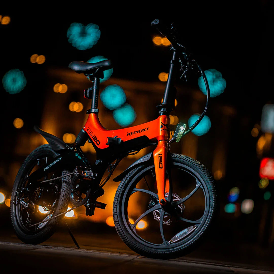 Izdelek je električno kolo, znamke MS Energy, model i20. Na sliki je vidno kolo v svetlo oranžni barvi, ki se nahaja v nočnem okolju s svetlimi ozadji. Ima manjša kolesa in športno obliko, kar nakazuje na njegovo uporabnost v urbanem okolju. Kolo je opremljeno z LED lučmi in ima sodoben dizajn.