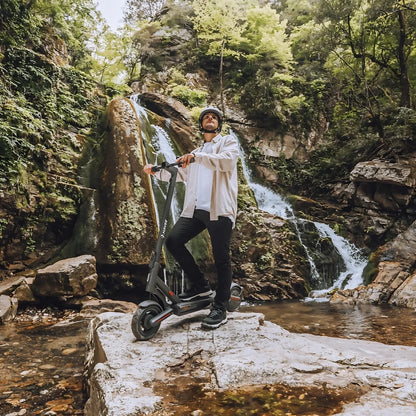 Oseba stoji na skali ob slapu, poleg električnega skiroja Navee S40.