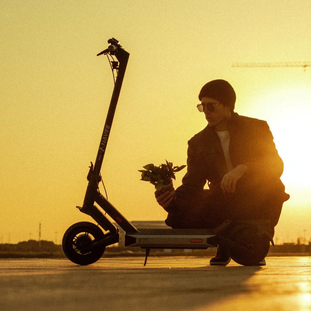 Oseba s sončnimi očali kleči ob električnem skiroju Navee S40 ob sončnem zahodu in drži rastline.