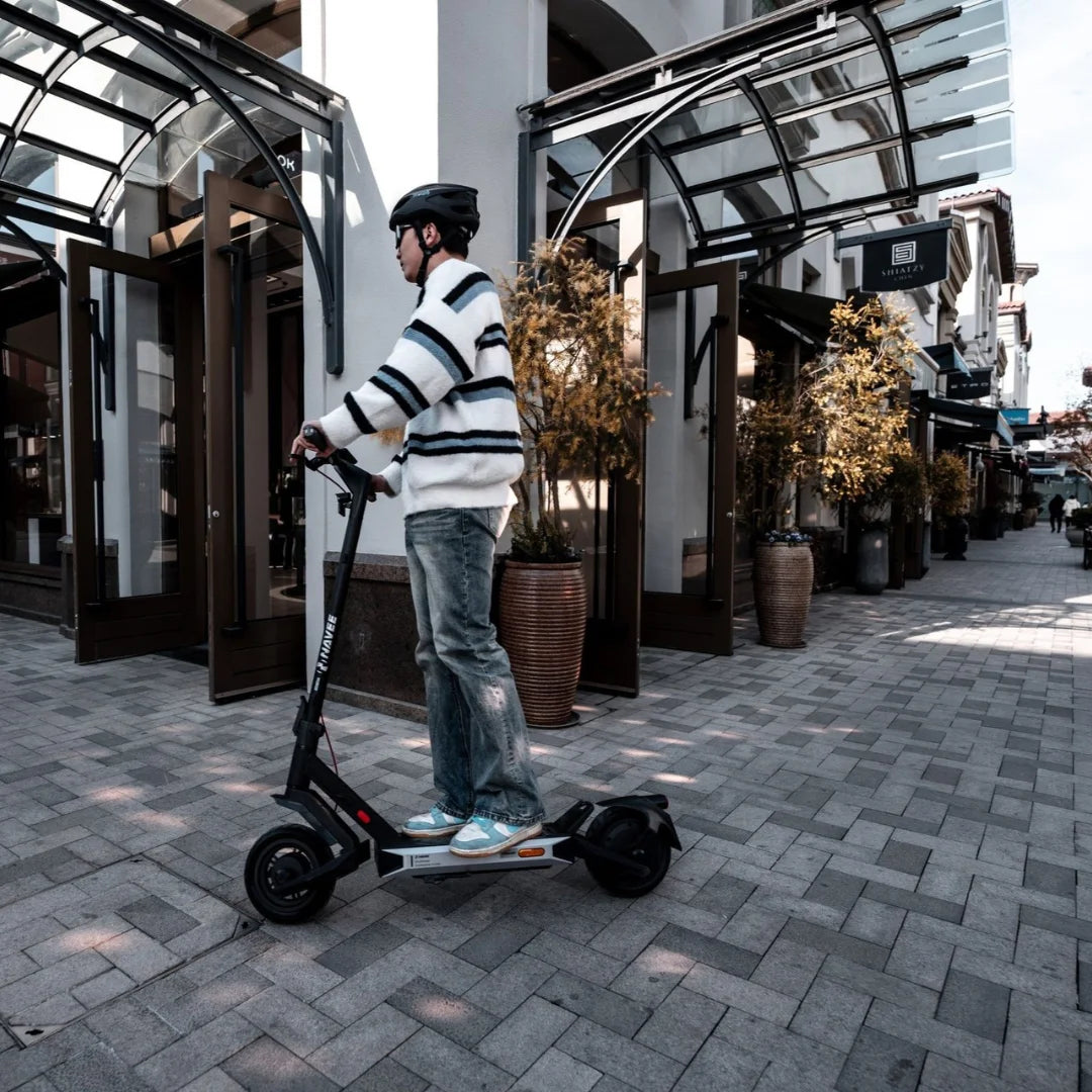 Oseba se vozi z električnim skirojem Navee S40 po tlakovani sprehajalni poti v bližini trgovin.