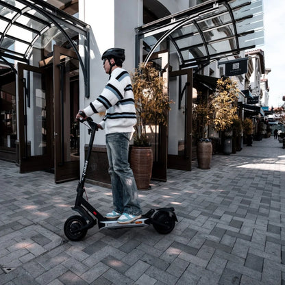 Oseba se vozi z električnim skirojem Navee S40 po tlakovani sprehajalni poti v bližini trgovin.