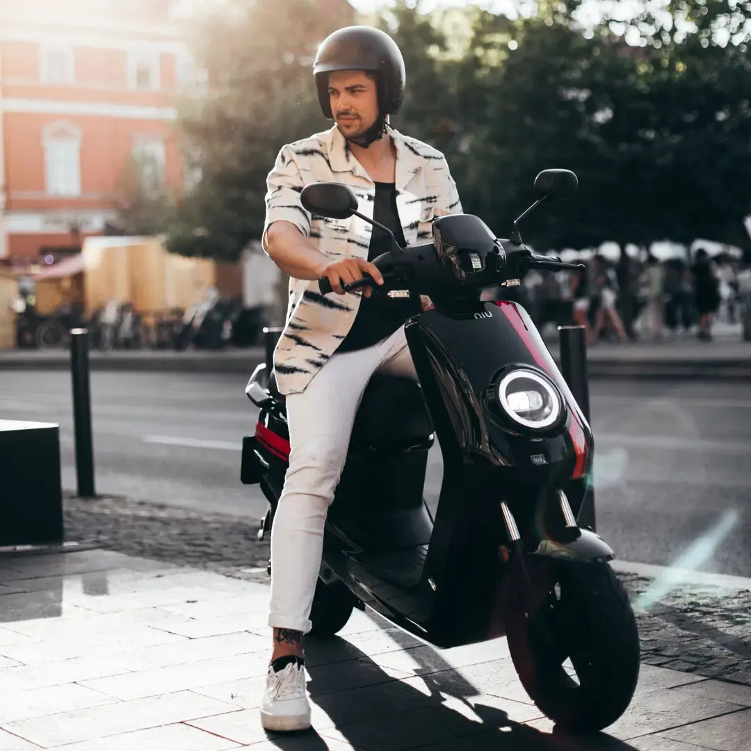 Moški s čelado sedi na električnem skuterju Niu Mqi Plus Sport v urbanem okolju.