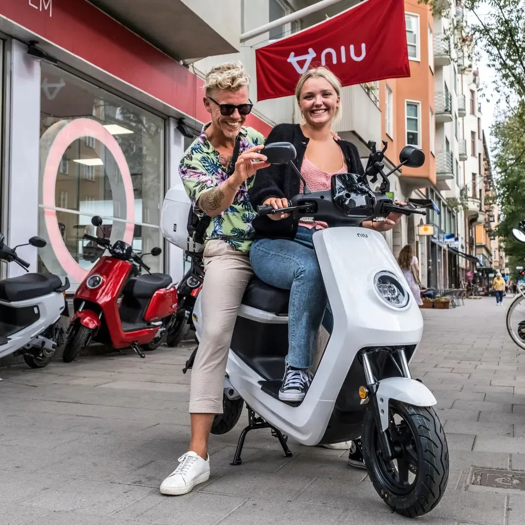 Par pozira na električnem skuterju Niu Mqi Plus Sport na živahni mestni ulici.
