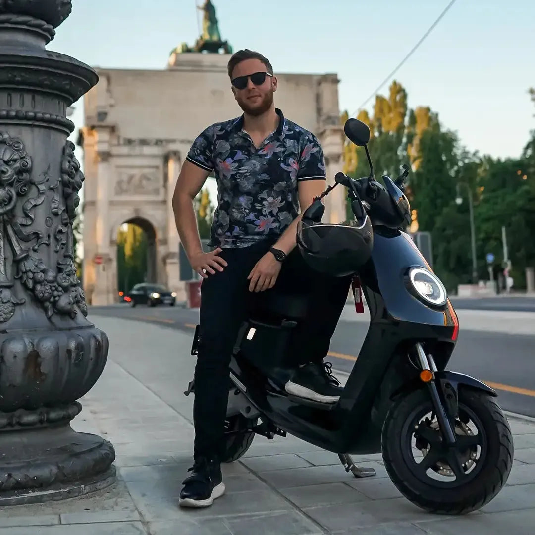 Moški s sončnimi očali se naslanja na električni skuter Niu Mqi Plus Sport blizu spomenika.