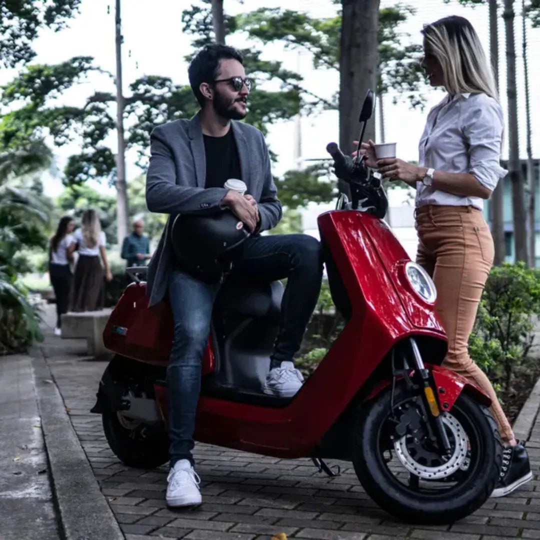 Moški sedi na rdečem električnem skuterju Niu Nqi, medtem ko ženska stoji v bližini, oba pa uživata v pijači.
