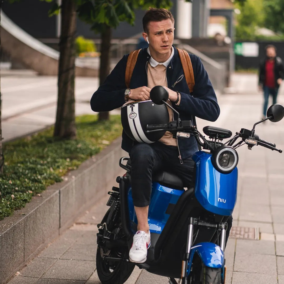 Moški sedi na električnem skuterju Niu Uqi GT in drži čelado v urbanem okolju.