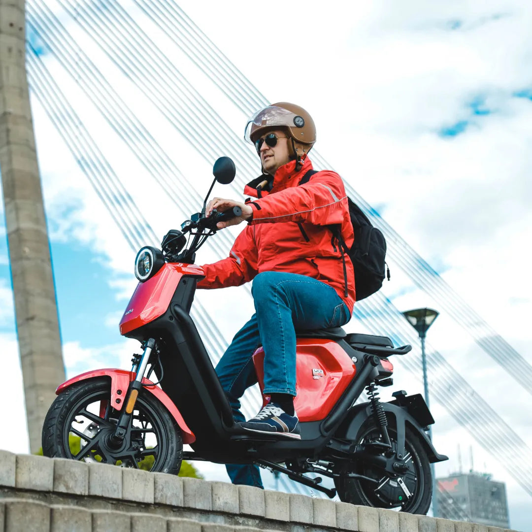 Oseba v rdeči jakni vozi električni skuter Niu Uqi GT v bližini mostu.