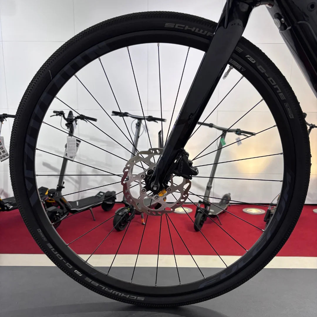 Izdelek na sliki je električno kolo, znamke Raymon, model GravelRay E 7.0. Na sliki je vidna sprednja kolesa s črnimi pnevmatikami in disk zavorami, kar nakazuje na športno zasnovo. V ozadju so vidna tudi električna skiroja, kar daje občutek o raznolikosti prevoznih sredstev v prostoru.