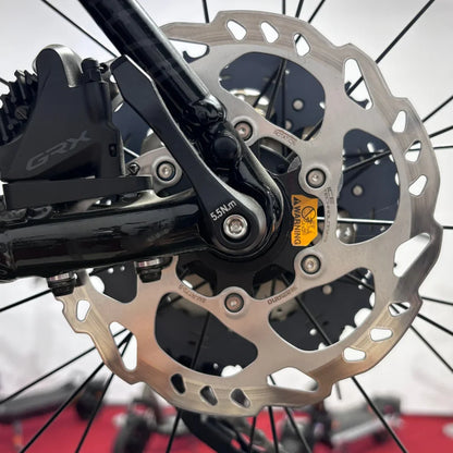 Izdelek je električno kolo, znamka je Raymon, model pa je GravelRay E 7.0. Na sliki je prikazan del zadnjega kolesa, kjer so vidni zavorni disk in sistem za prestavljanje. Zavorni disk je izdelan iz nerjavečega jekla, kar zagotavlja dobro zmogljivost pri zaviranju, medtem ko je na okvirju vidna oznaka za pravilno namestitev.