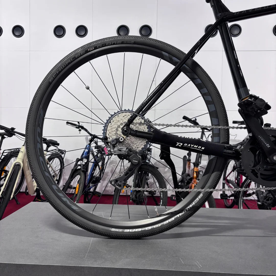 Izdelek na sliki je električno kolo, model Raymon GravelRay E 7.0. Na sliki je vidna zadnja kolesa s pnevmatikami primerne za gravel terene, ter del pogonskega sistema, vključno s kaseto in verigo. Kolo je postavljeno na stojalo, v ozadju pa so vidna še druga kolesa.