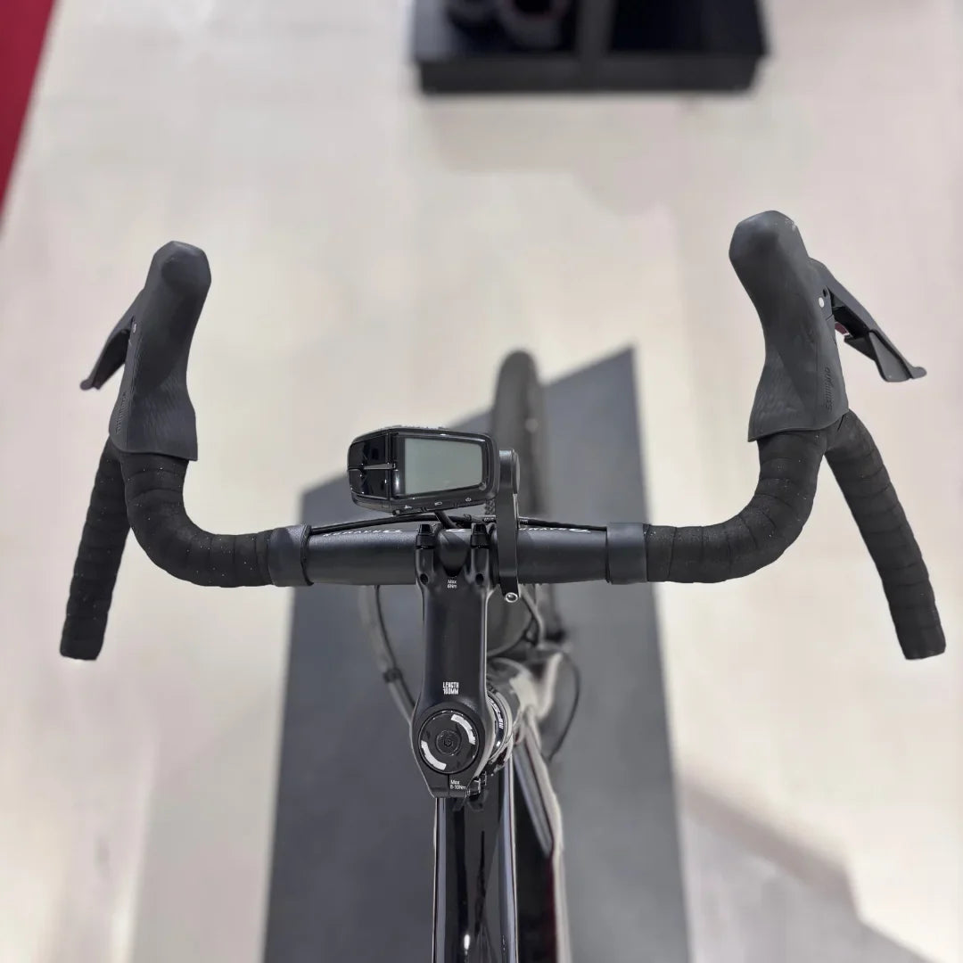 Izdelek je električno kolo, znamke Raymon, model GravelRay E 7.0. Na sliki je vidna sprednja stran krmila, ki ima športne ročke in digitalni zaslon, verjetno za prikaz informacij o vožnji. Kolo ima tudi elegantno črno barvo in je zasnovano za gravel vožnjo.