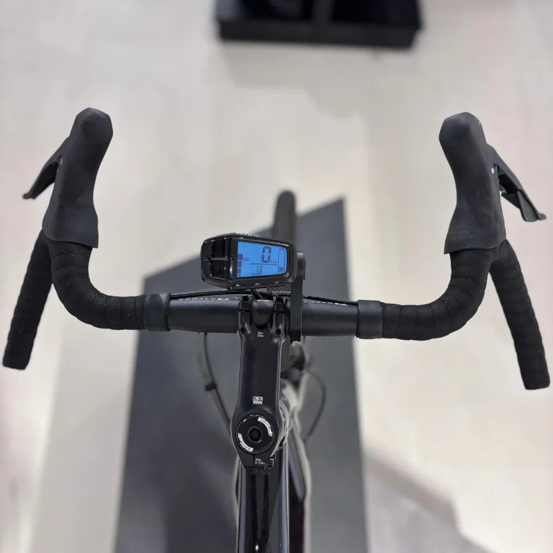 Izdelek na sliki je električno kolo, model Raymon GravelRay E 7.0. Na sliki so vidni krmili s prevleko iz gume ter digitalni zaslon, ki prikazuje hitrost in druge podatke. Kolo je zasnovano za gravel vožnjo, kar pomeni, da je primerno za različne terene.