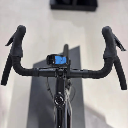 Izdelek na sliki je električno kolo, model Raymon GravelRay E 7.0. Na sliki so vidni krmili s prevleko iz gume ter digitalni zaslon, ki prikazuje hitrost in druge podatke. Kolo je zasnovano za gravel vožnjo, kar pomeni, da je primerno za različne terene.