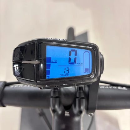 Izdelek je električno kolo, znamke Raymon, model GravelRay E 7.0. Na sliki je prikazan digitalni zaslon, ki prikazuje hitrost 0 km/h ter razdaljo 13 km, kar nakazuje preostalo razdaljo, ki jo lahko prevozite. Zaslon je nameščen na krmilu kolesa.