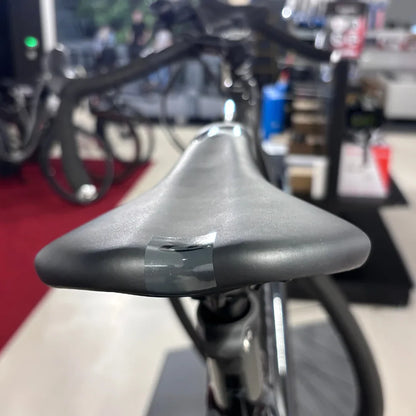 Izdelek na sliki je električno kolo, znamke Raymon, model GravelRay E 7.0. Na sliki je prikazan sedež kolesa, ki je črn in ima na sebi lepilni trak, kar nakazuje na morebitno poškodbo ali popravilo. V ozadju se vidi del kolesa in okolica trgovine.