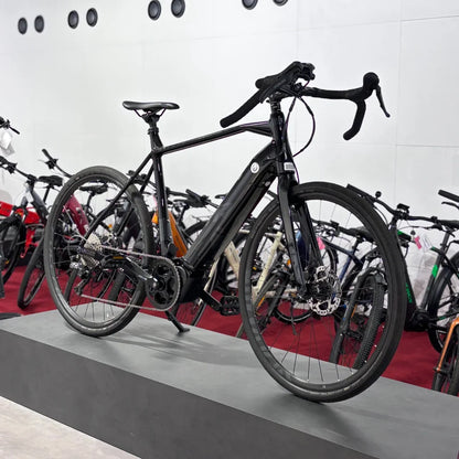 Izdelek na sliki je električno kolo, znamke Raymon, model GravelRay E 7.0. Kolo je črne barve in ima športno obliko, kar je značilno za gravel kolesa. Na sliki je postavljeno na podstavek, v ozadju pa so vidna še druga kolesa. Kolo ima široke gume in disk zavore, kar omogoča dobro oprijemljivost in zaviranje.