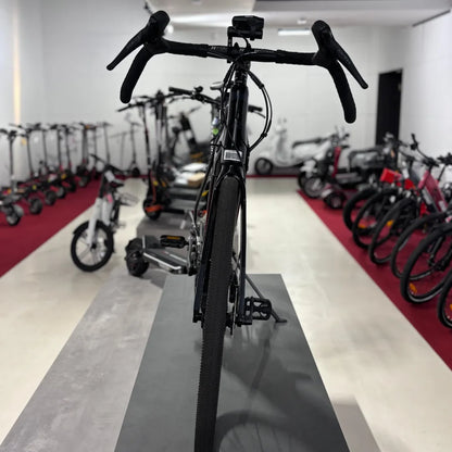 Izdelek na sliki je električno kolo, natančneje model Raymon GravelRay E 7.0. Na sliki je vidna zadnja stran kolesa z vidnimi krmili in širokimi pnevmatikami, kar nakazuje na njegovo primernost za gravel terene. V ozadju so tudi drugi električni skiroji in kolesa, kar kaže na prodajno okolje.