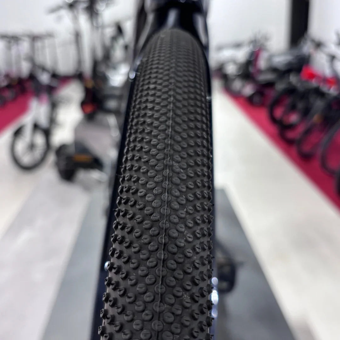 Izdelek na sliki je električno kolo, znamke Raymon, model GravelRay E 7.0. Na sliki je viden profil kolesa s posebno teksturo pnevmatike, ki je zasnovana za vožnjo po različnih terenih. V ozadju so vidne druge kolesarske naprave, kar nakazuje, da je slika posneta v trgovini ali razstavi.