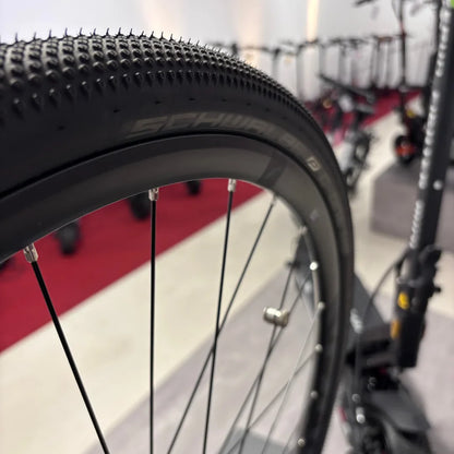 Izdelek je električno kolo, znamka je Raymon, model pa GravelRay E 7.0. Na sliki je prikazan del kolesa, natančneje pnevmatika s profilom, ki je primeren za vožnjo po gravel površinah, ter del obroča in žice. V ozadju so vidni tudi drugi kolesi, kar nakazuje, da gre za razstavo ali trgovino.